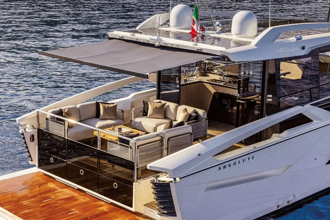 Luxury yacht with modern seating area, grey awning, and an Italian flag, alongside calm waters and wooden deck.
